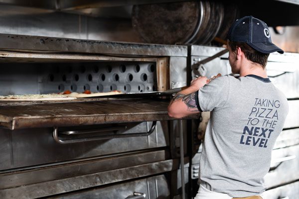 A man in a Dewmore charity shirt uses a wooden paddle to remove a pizza from the oven