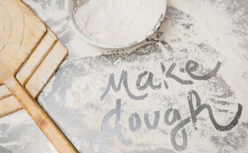 The words "make dough" spelled out in flour dusted on a table. A pizza peel lays near by.