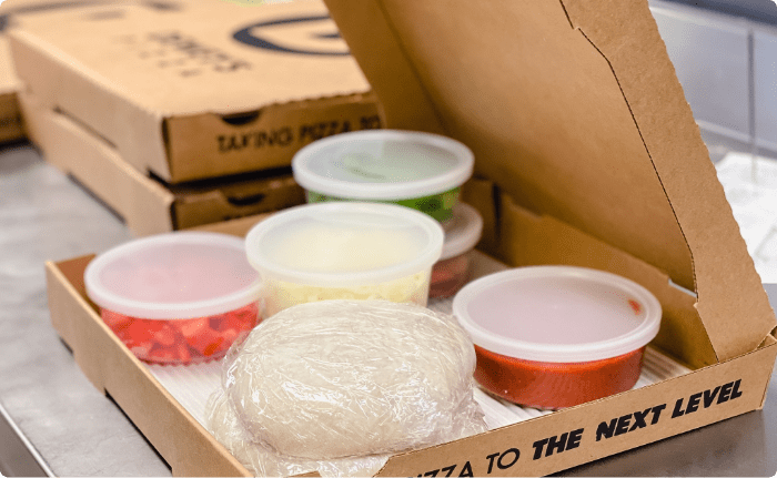 an open pizza box containing several plastic food containers of individual pizza ingredient