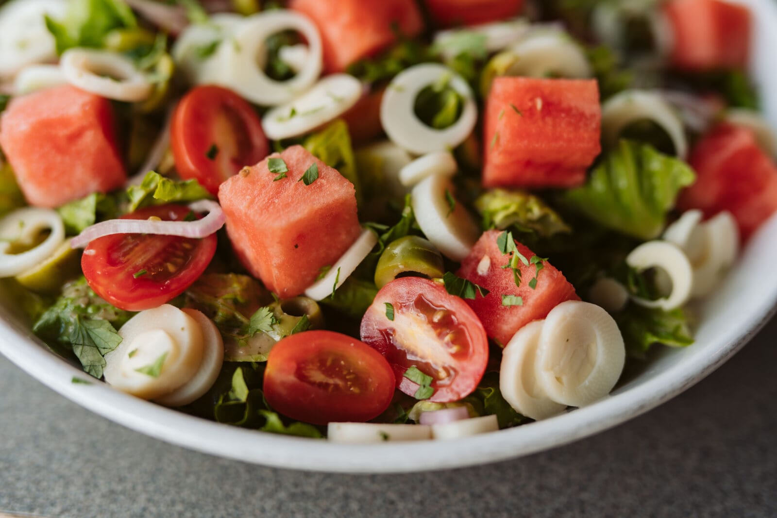 A vibrant close-up of the Palmitas seasonal salad served in a white bowl. Crisp romaine lettuce is tossed with bright red cubes of watermelon, juicy halved cherry tomatoes, sliced white rings of hearts of palm, and green Castelvetrano olives, all garnished with finely chopped fresh herbs and thin shallot slivers.