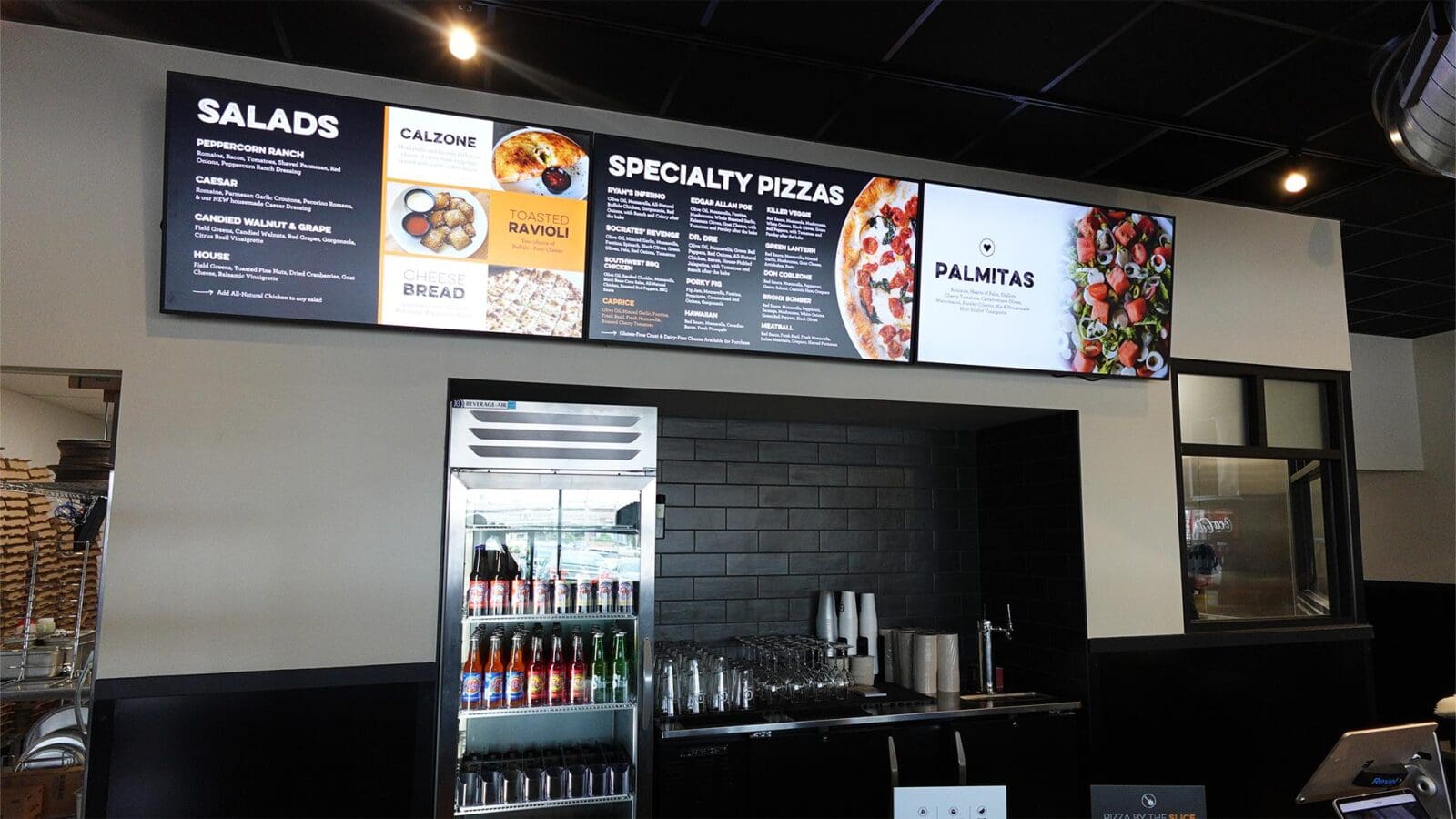 Digital menu boards mounted on a wall inside Dewey's Pizza in Creve Coeur, showing categories for salads, calzones, specialty pizzas, and seasonal promotions above a glass beverage cooler.