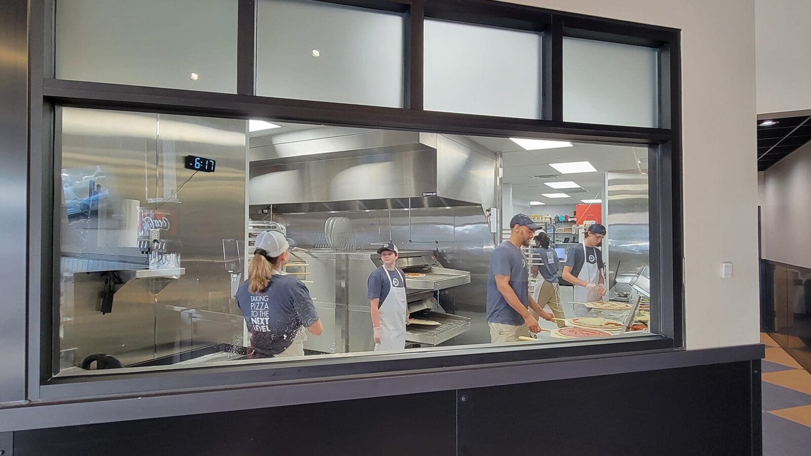View through the dough toss window showing kitchen staff inside Dewey's Pizza in Creve Coeur preparing and assembling pizzas.
