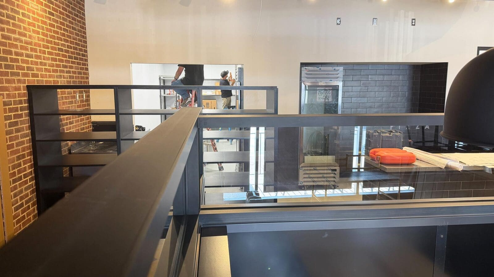 An interior view of the new Dewey's Pizza location in Creve Coeur under construction, looking past dark gray dividing walls and shelving toward a back room where construction workers are active on ladders.