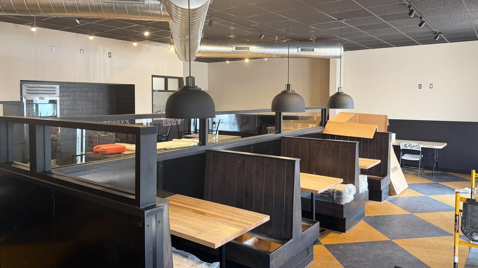 The main dining area of the new Dewey's Pizza in Creve Coeur during construction, showing a row of dark wood booths with light wood tables underneath hanging black dome pendant lights.