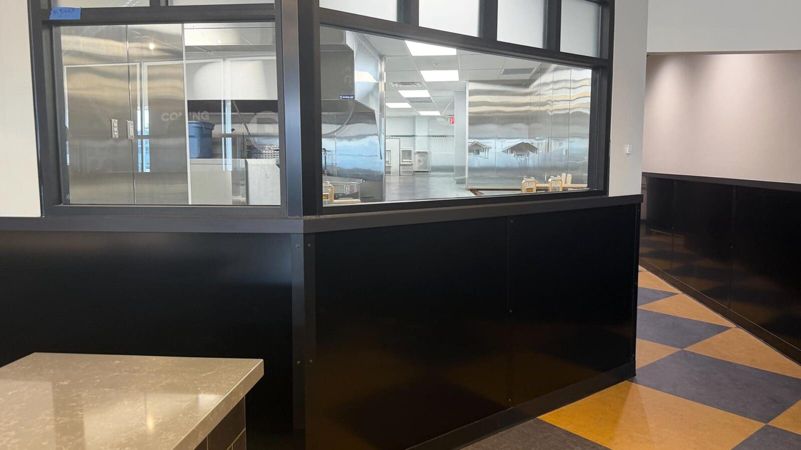 An interior view of the new Dewey's Pizza location in Creve Coeur, featuring a sleek black partition wall with a large glass dough tossing window looking into a clean, stainless steel kitchen.