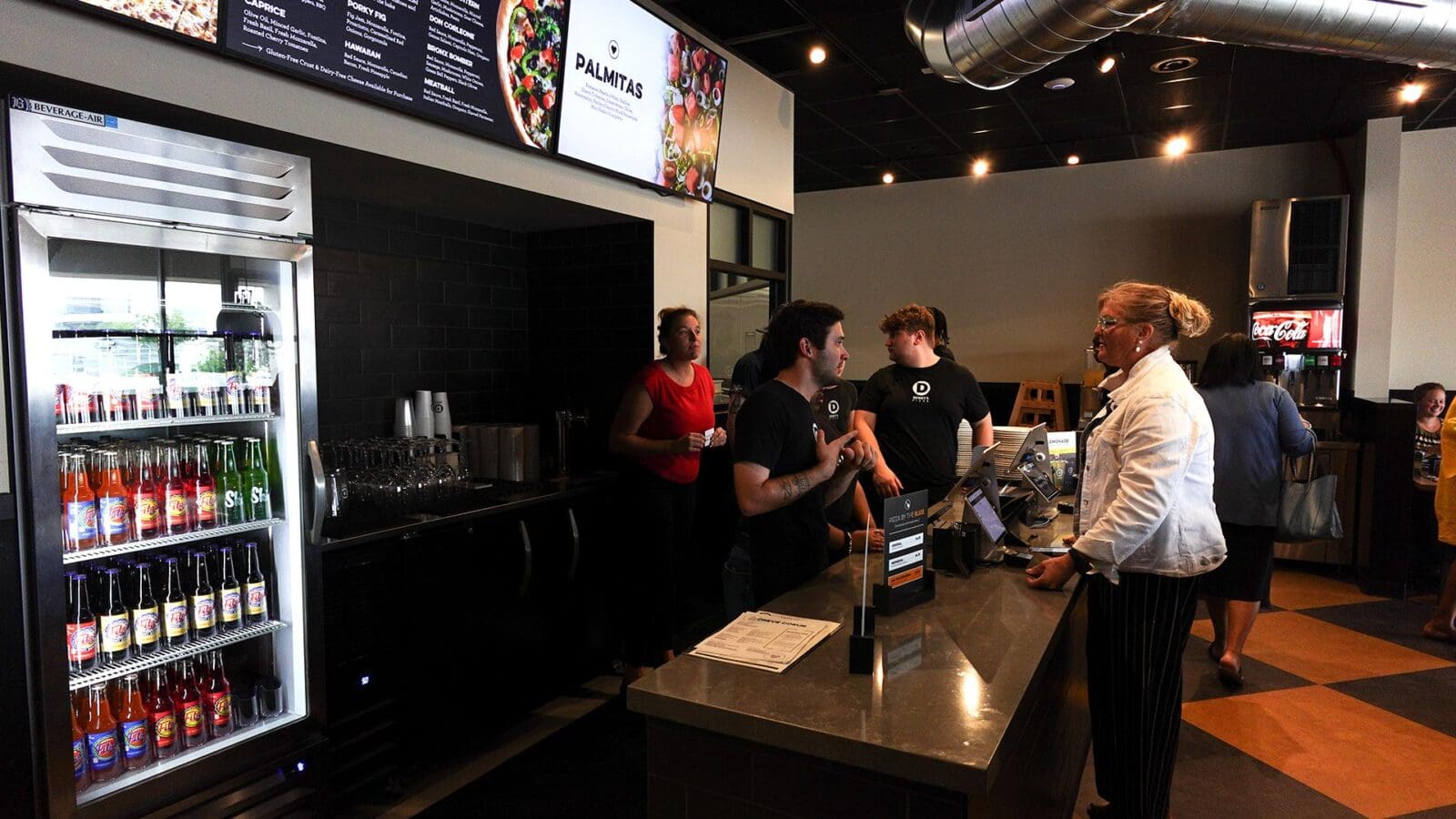 Interior view of Dewey's Pizza counter in Creve Coeur, featuring employees taking customer orders, digital menu boards overhead, and a glass-door beverage cooler stocked with bottled drinks.