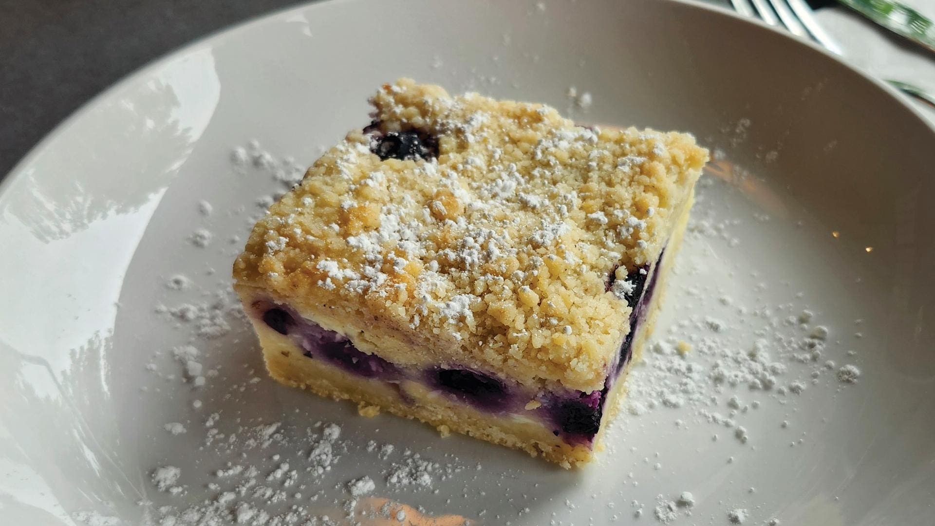 A square lemon blueberry bar with a crumb topping and a dusting of powdered sugar, served on a white plate.
