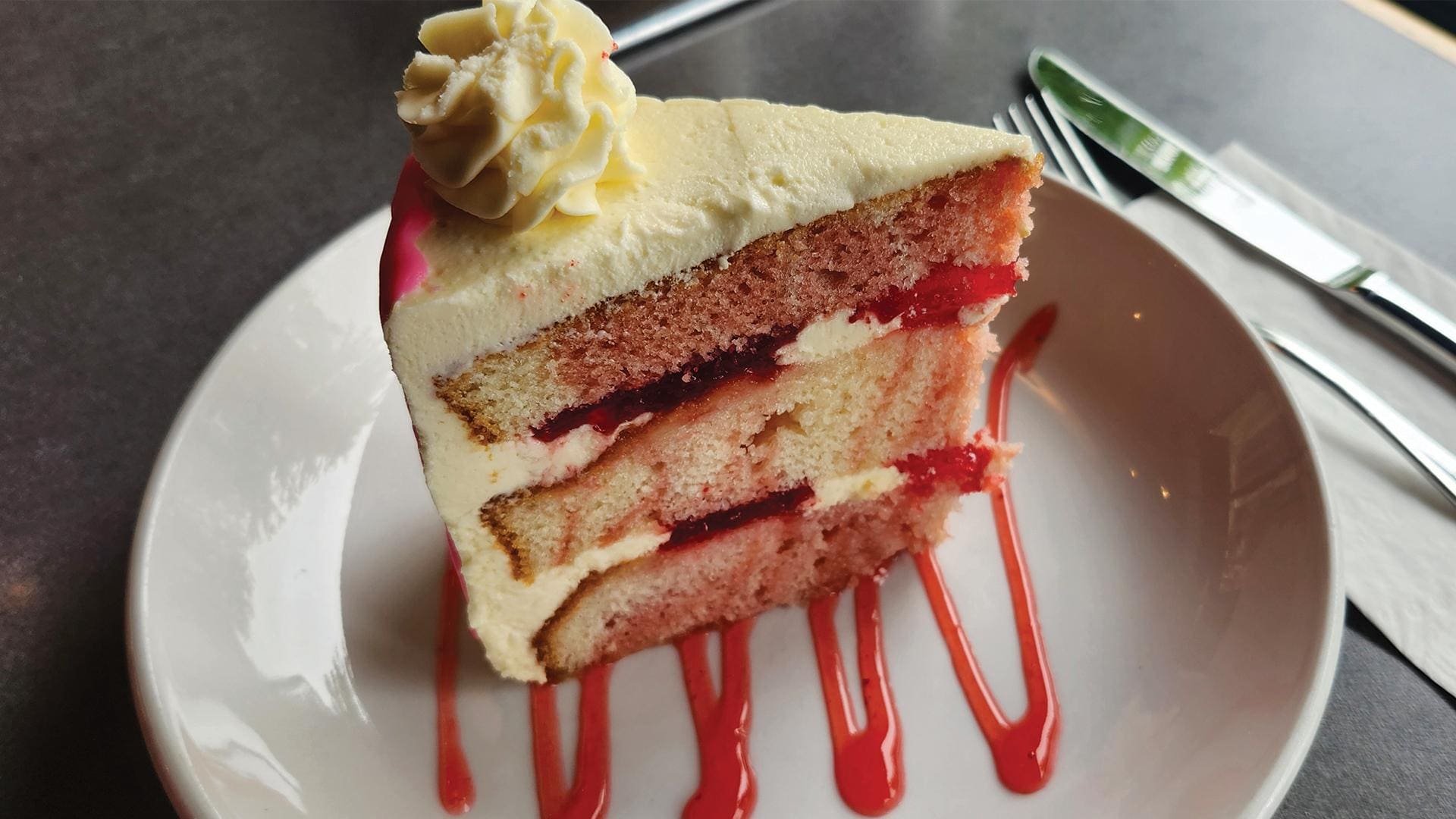 A slice of strawberry swirl layer cake with white frosting, a dollop of whipped cream on top, and a drizzle of red strawberry sauce on the white plate.