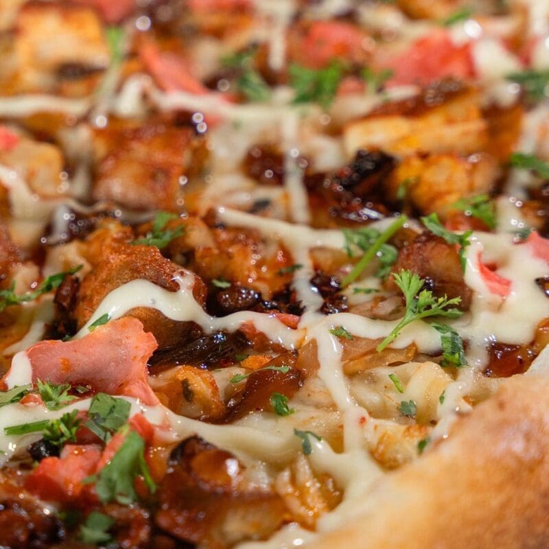 A close-up, top-down view of a Dewey’s Korean BBQ seasonal pizza. The pizza features our ingredients which include Sweet Chili Sauce, Mozzarella, Kimchi, Soy Brown Sugar Caramelized Onions, Diced Korean BBQ Chicken Thighs, Pickled Ginger, with Kewpie Mayo, Chili Crisp and Parsley & Cilantro mix after the bake. The crust is thick, golden, and perfectly toasted.