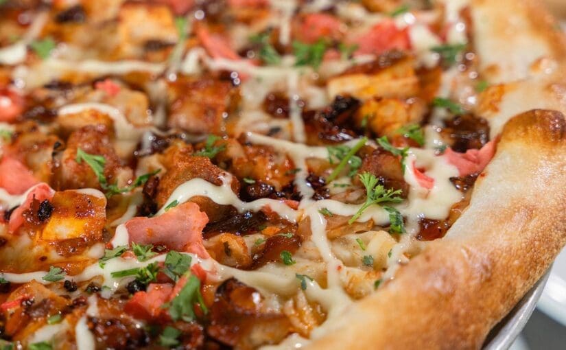 A close-up, top-down view of a Dewey’s Korean BBQ seasonal pizza. The pizza features our ingredients which include Sweet Chili Sauce, Mozzarella, Kimchi, Soy Brown Sugar Caramelized Onions, Diced Korean BBQ Chicken Thighs, Pickled Ginger, with Kewpie Mayo, Chili Crisp and Parsley & Cilantro mix after the bake. The crust is thick, golden, and perfectly toasted.