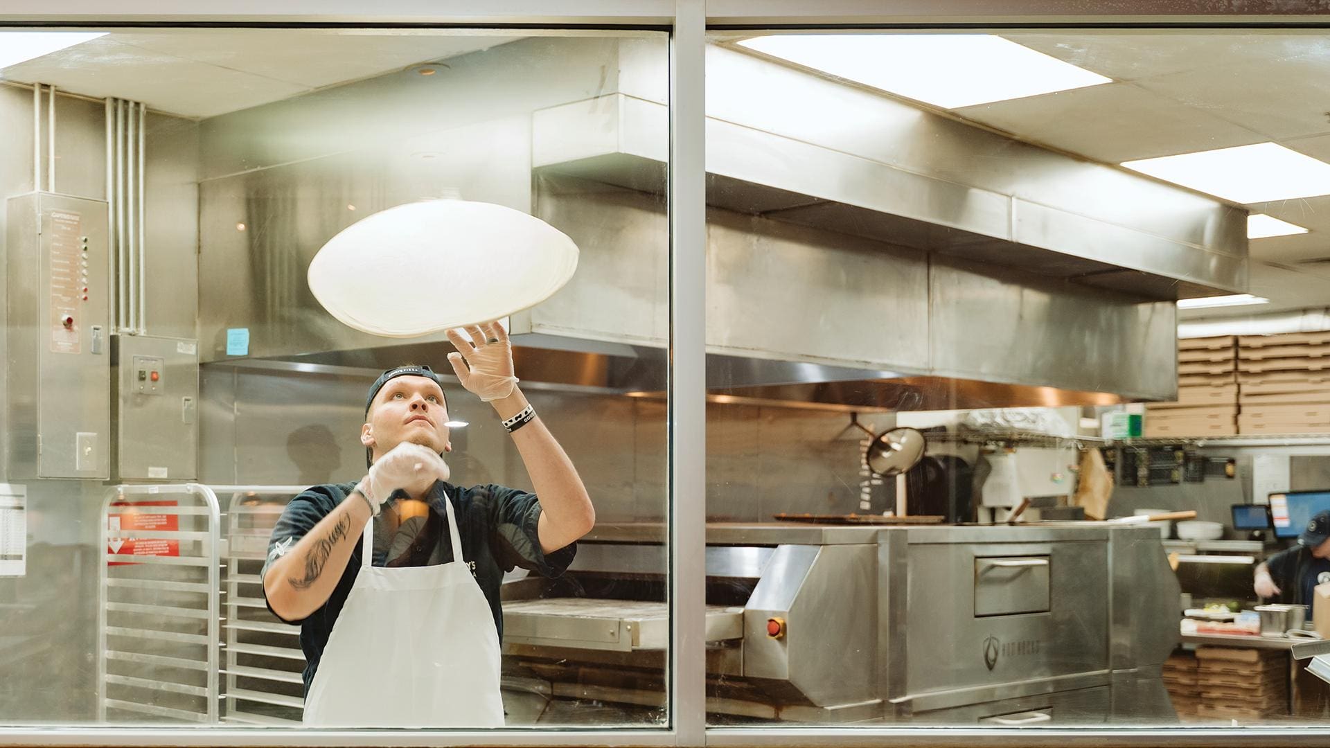 A pizza maker wearing a black cap, black shirt, and a white apron stands in a commercial kitchen, looking upward as he tosses a round, spinning pizza dough high into the air. He wears clear plastic gloves, and a tattoo is visible on his right forearm. The scene is viewed through a large glass window, showing the stainless steel kitchen equipment, including a pizza oven, ventilation hoods, and stacks of pizza boxes in the background.