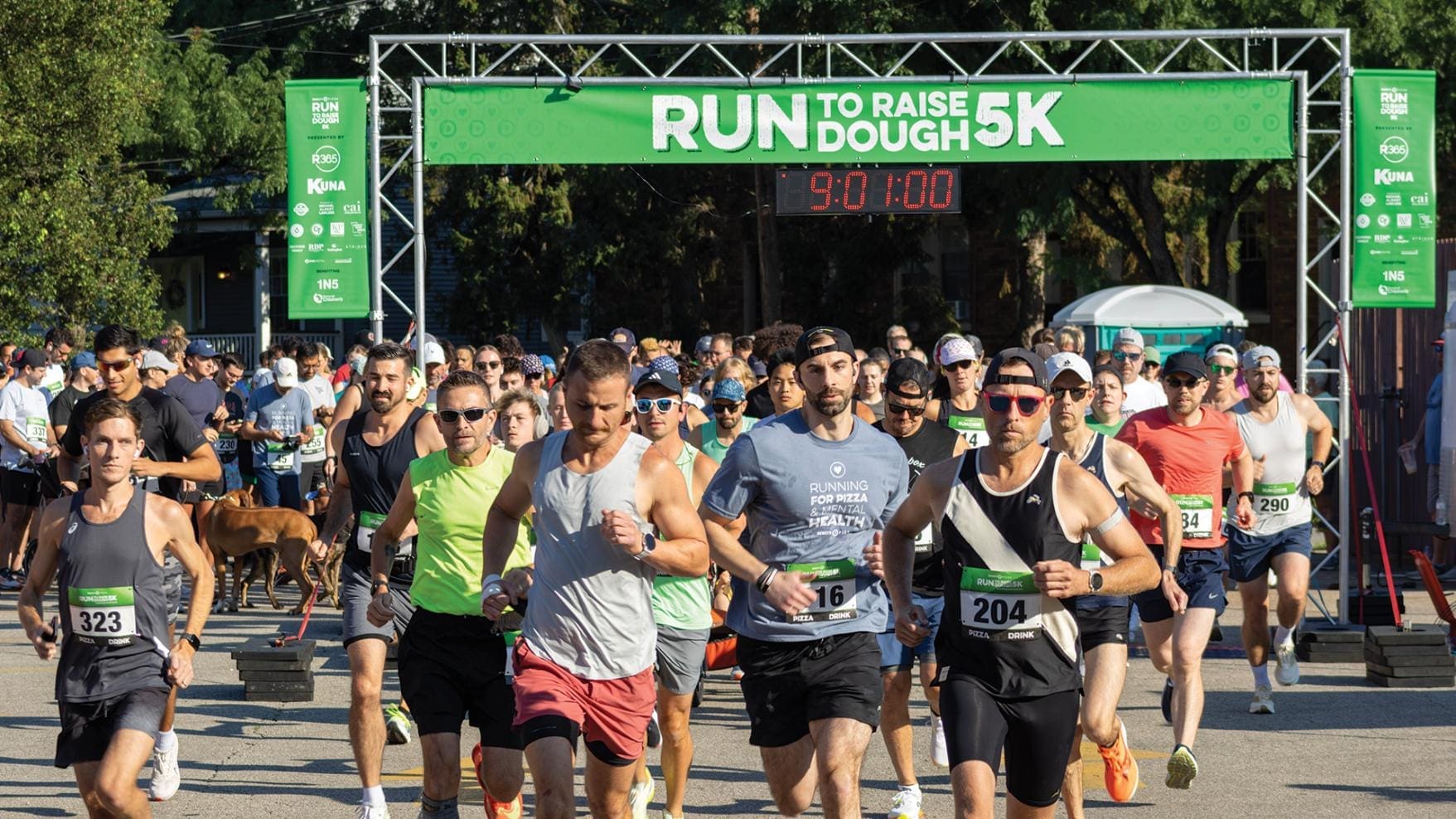 A large crowd of runners crossing the starting line of the "Run to Raise Dough 5K" under a green banner and a digital race clock reading 9:01:00. The scene is sunny and high-energy, featuring participants in colorful athletic wear.