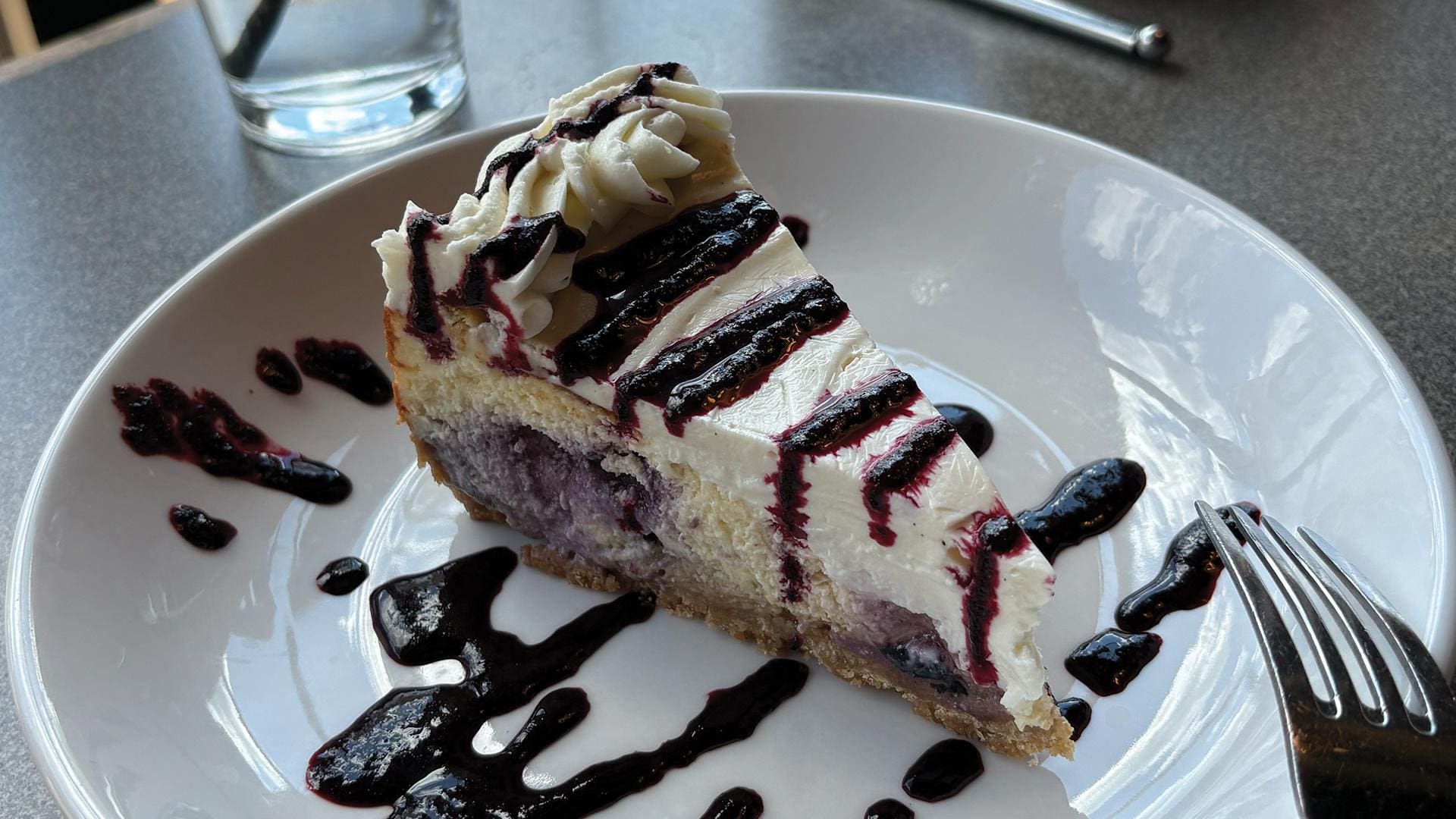 A slice of seasonal lemon blueberry cheesecake on a white plate, featuring a creamy lemon filling loaded with sweet blueberries on a graham cracker crust, topped with a smooth sour cream layer and a bright drizzle of blueberry purée.
