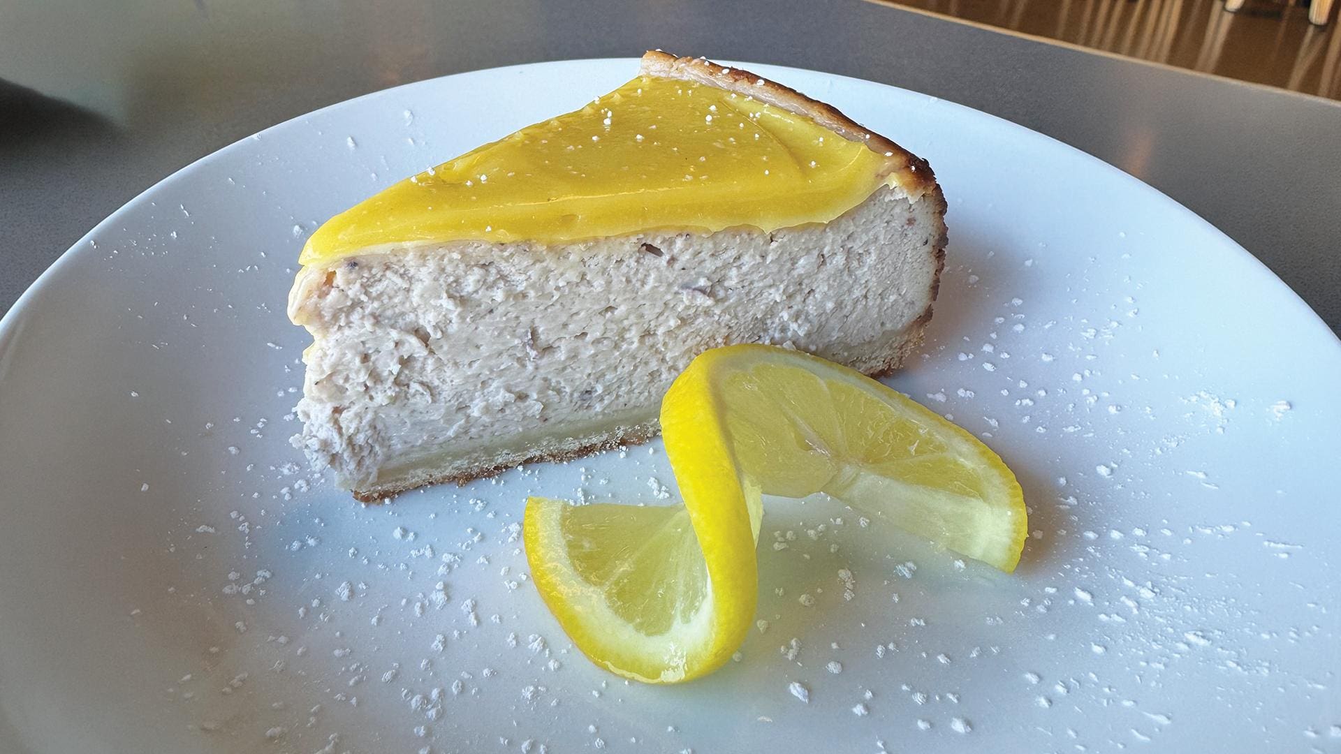 A slice of lemon blueberry cheesecake on a white plate, topped with a bright yellow lemon curd and served with a twisted lemon slice.