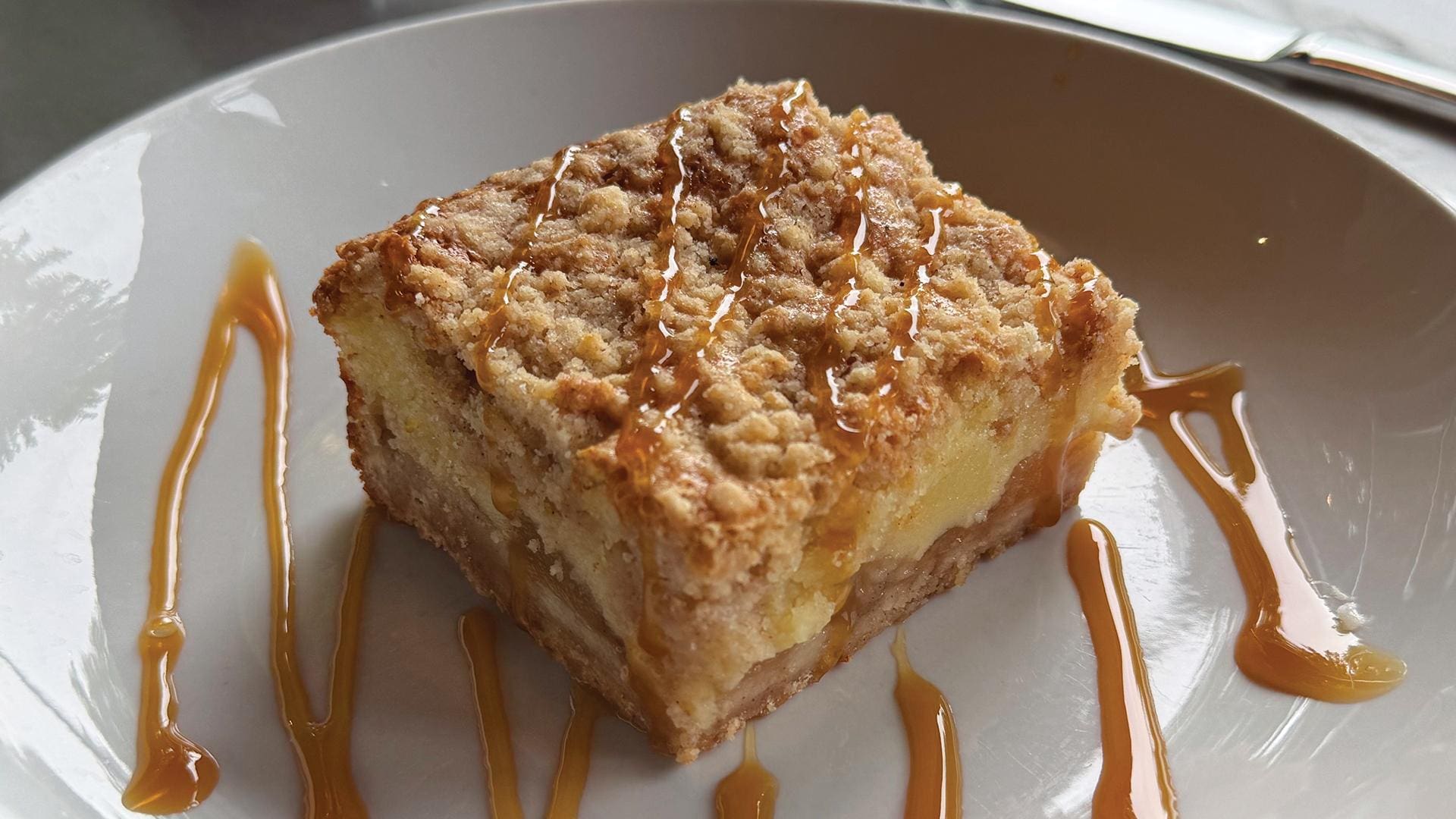 A close-up shot of a square apple pie bar served on a white plate. The dessert features a distinct layered look with a soft crust bottom, a thick center layer of sweet baked apples, and a golden-brown, textured cobbler crumb topping scattered with crystallized sugar. The top of the bar and the surrounding plate are decorated with a neat drizzle of glossy, golden caramel sauce.