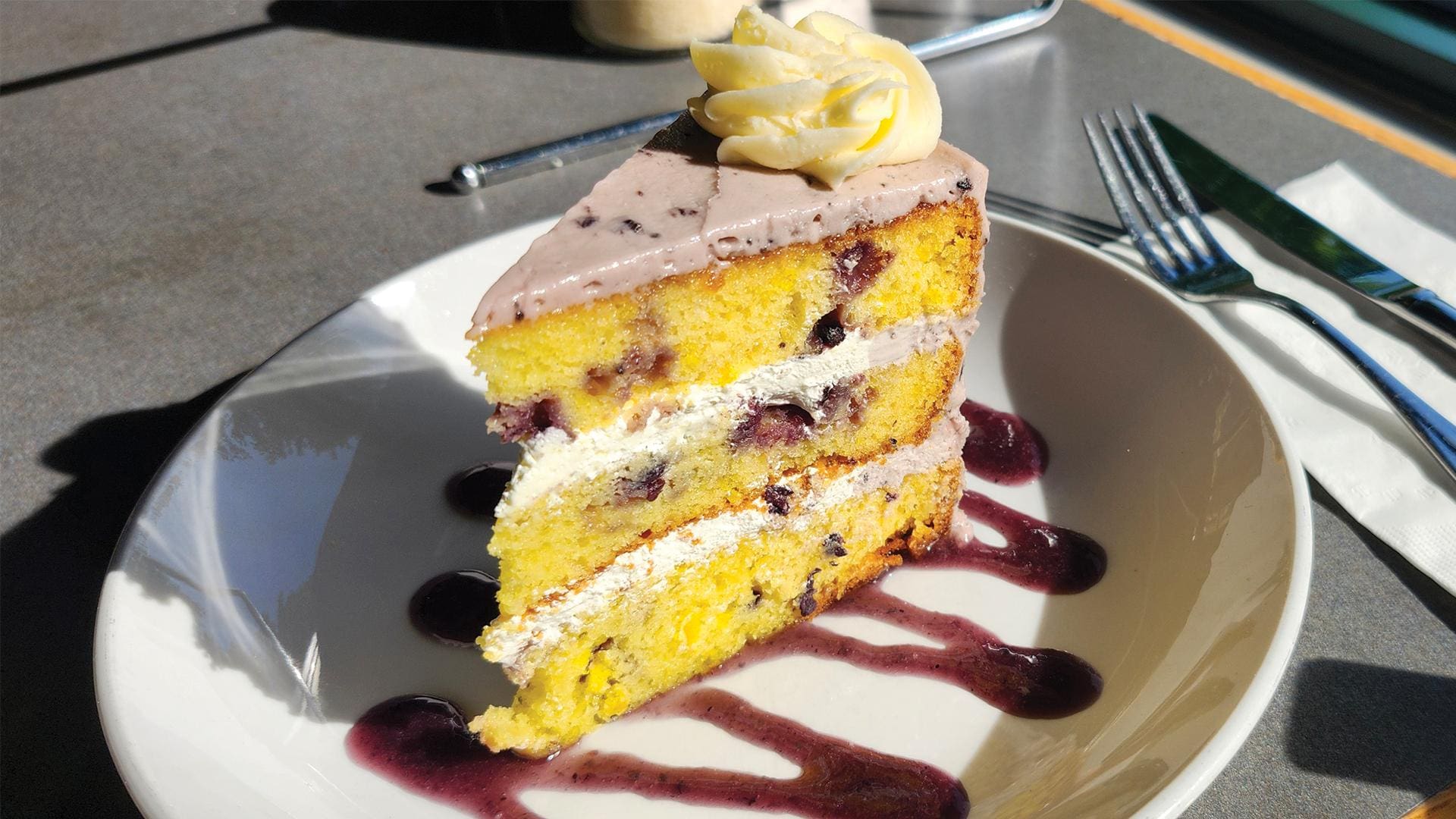 A slice of a triple-layer lemon blueberry cake on a white plate, illuminated by bright sunlight. The cake features bright yellow sponge layers packed with whole blueberries, separated by a light lemon mousse filling. It is topped with pale purple blueberry buttercream frosting, a classic yellow buttercream rosette, and sits over a zig-zag drizzle of vibrant blueberry puree. A fork and knife rest on a napkin in the background.