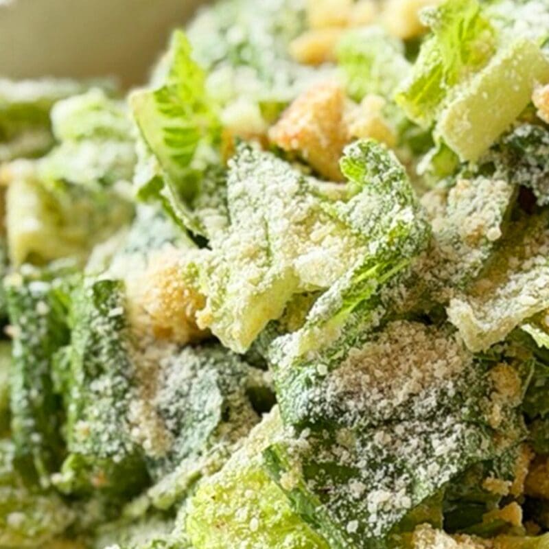 Freshly tossed Caesar salad in a white bowl from Dewey's Pizza, featuring Romaine, Parmesan Garlic Croutons, Pecorino Romano, and our new housemade Caesar Dressing