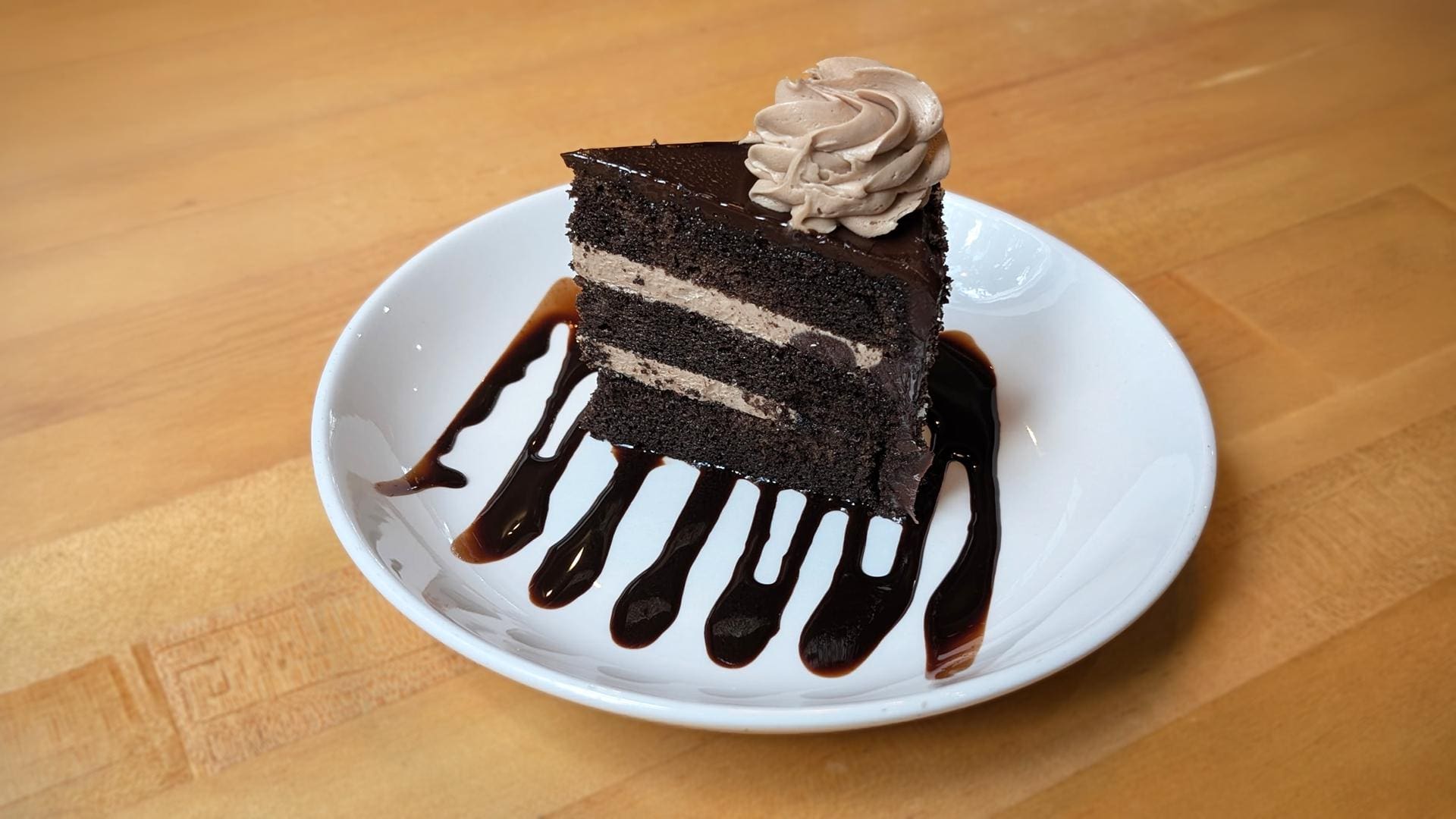 A slice of three-layer chocolate blackout cake on a white plate, drizzled with chocolate sauce and topped with a swirl of light brown frosting, set on a light wood table.
