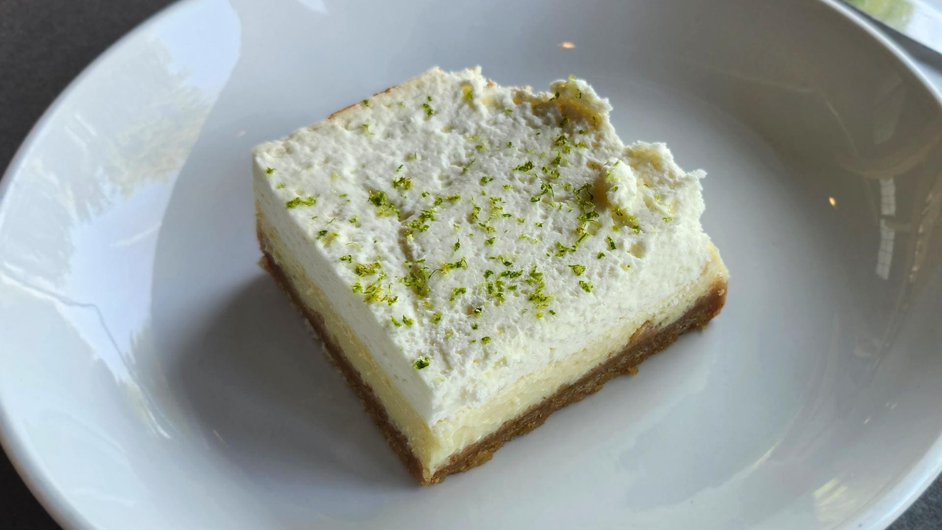 A square key lime pie bar on a white plate with a buttery graham cracker crust, tangy key lime filling, soft whipped cream topping, and fresh lime zest.