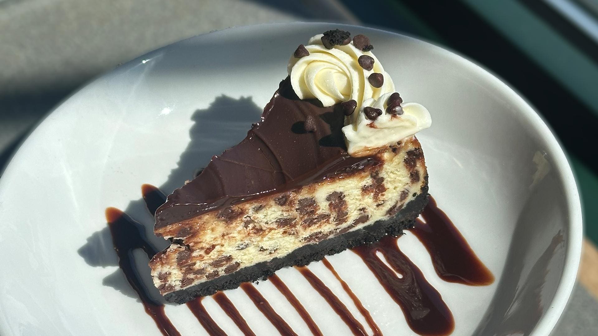 A slice of chocolate chip cheesecake with a dark chocolate crust and smooth ganache topping, served on a white plate with chocolate drizzle.