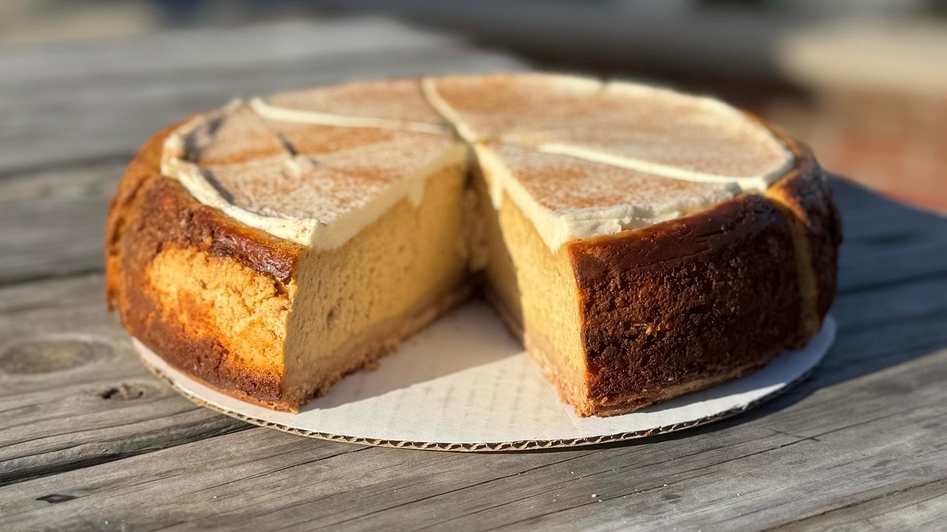 Whole pumpkin cheesecake from The Cheesecake Girl on a cardboard cake board, sitting on a wooden outdoor table with a slice removed to reveal its thick filling.
