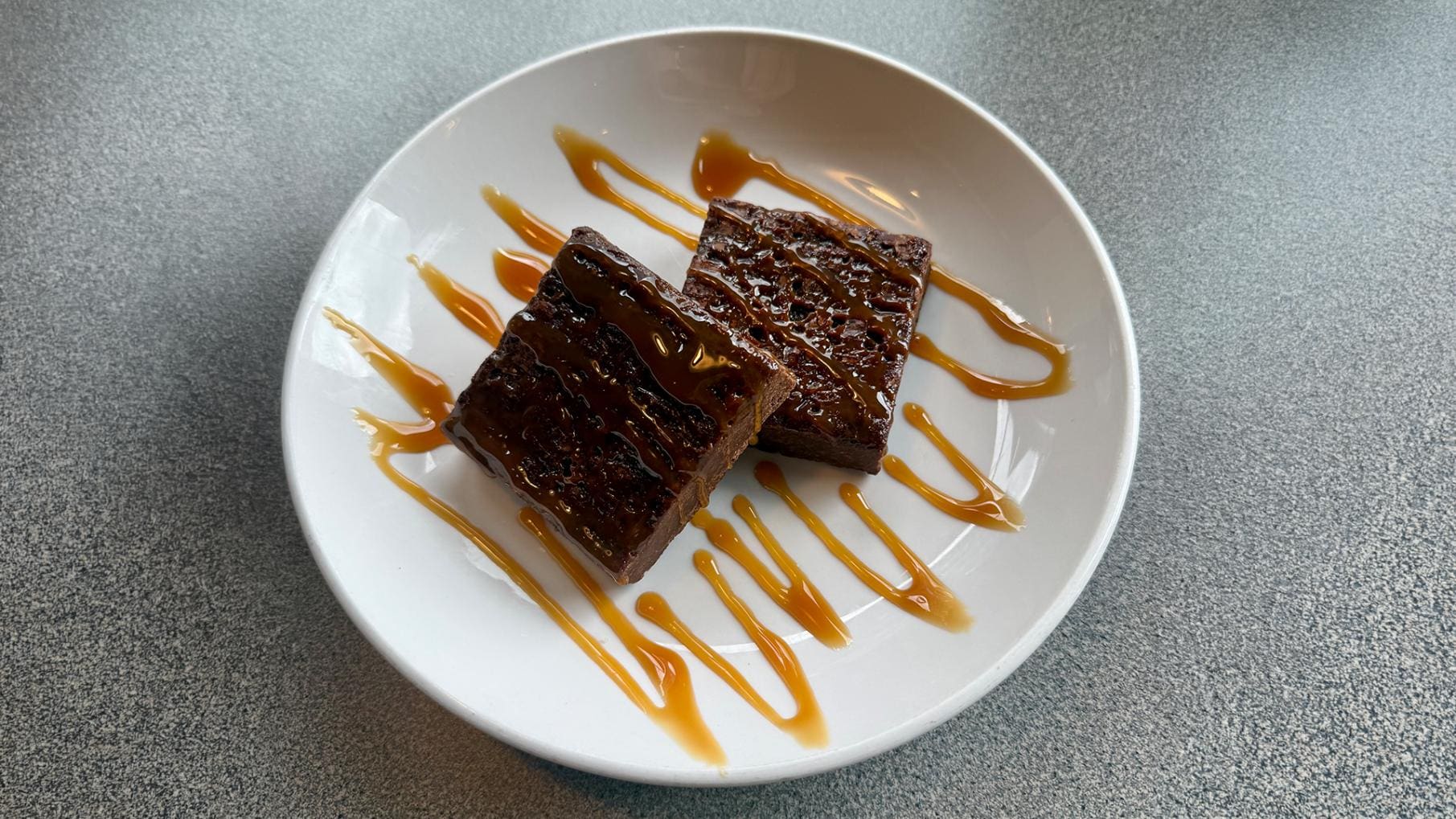Two Brownie Points fudge brownies drizzled with caramel sauce, served in a shallow white bowl on a gray table at Dewey's Pizza.