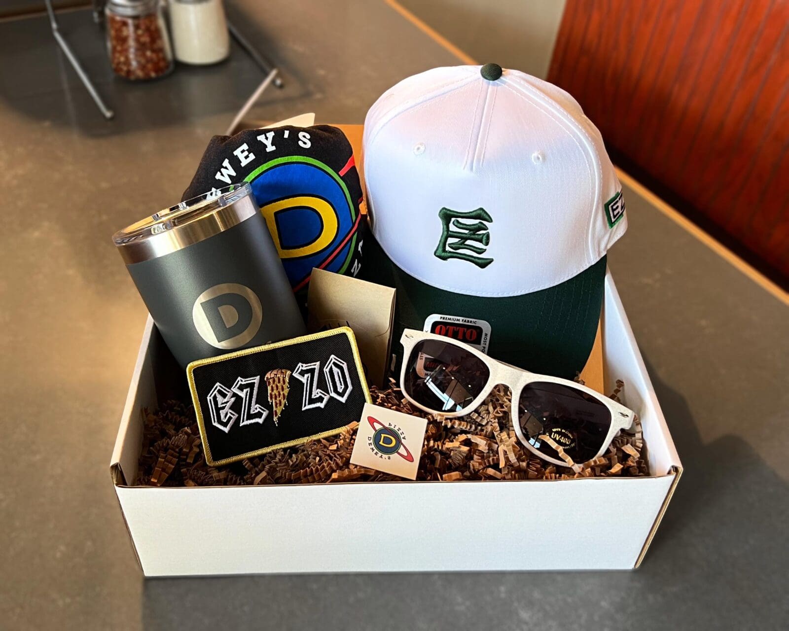 A white gift box filled with Dewey’s Pizza merchandise, including a white and green baseball cap, a dark grey tumbler with a 