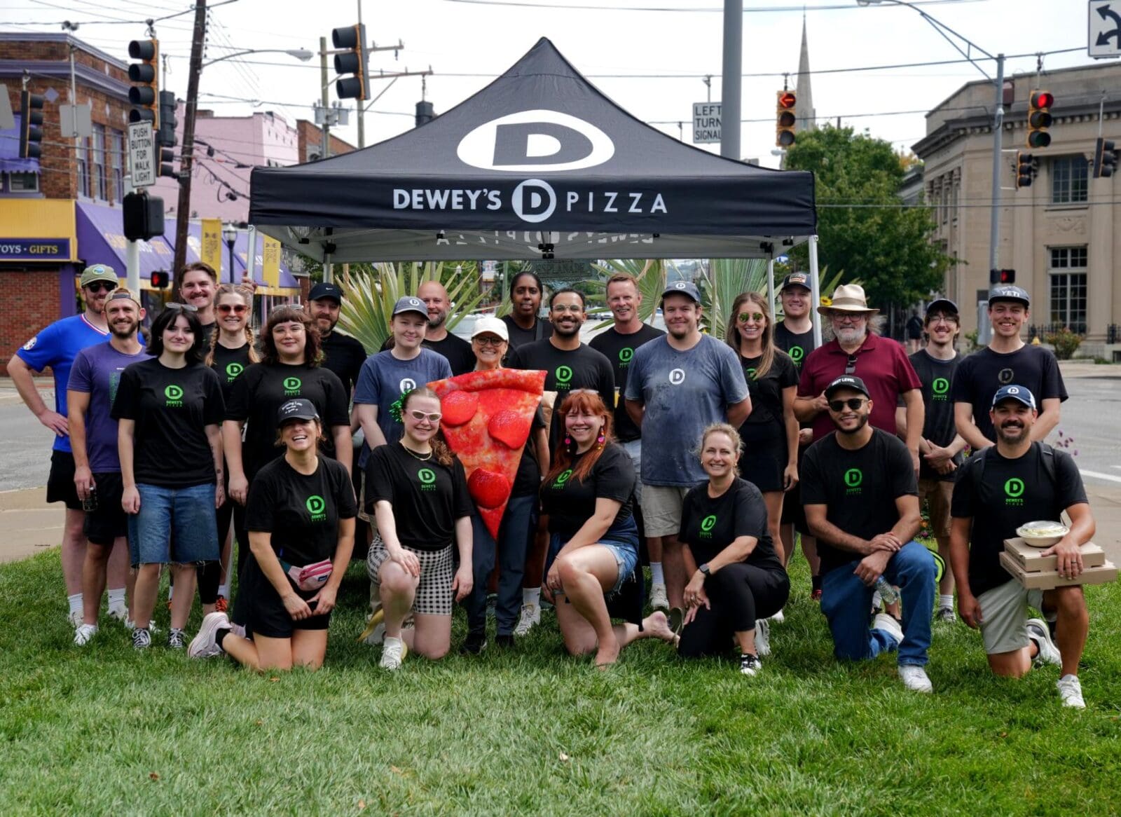 The Dewey's Pizza team posing together outdoors under a branded pop-up tent at the Run to Raise Dough 5K, with someone in the center wearing a large pepperoni pizza costume.