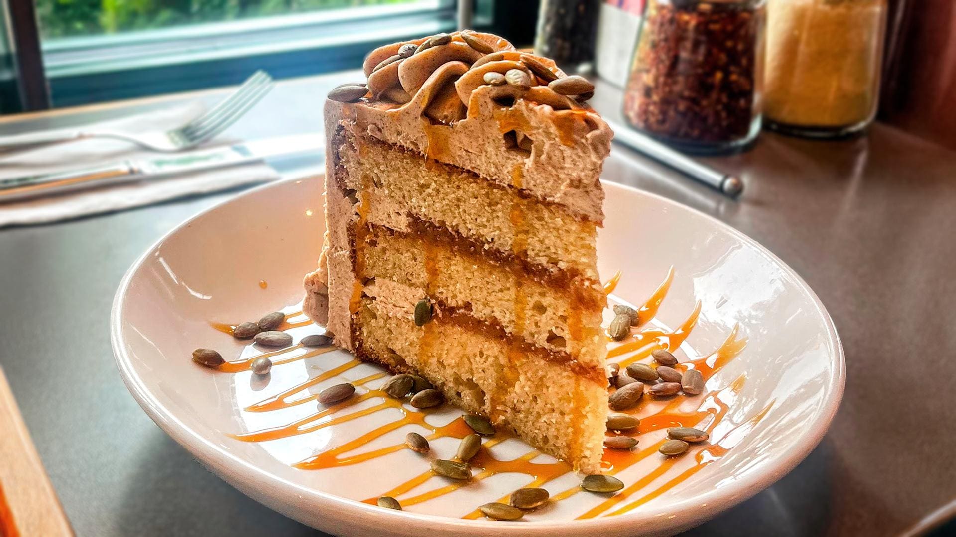A slice of three-layer caramel apple cider cake served on a white plate, topped with cinnamon buttercream, roasted pumpkin seeds, and a drizzle of caramel sauce.