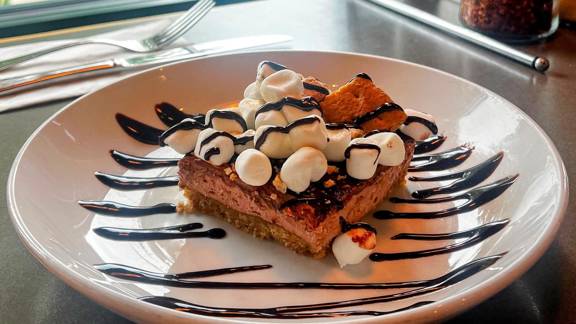 A French silk s’mores dessert bar on a graham cracker crust, piled high with mini marshmallows, graham cracker pieces, and drizzled with chocolate sauce on a white plate.